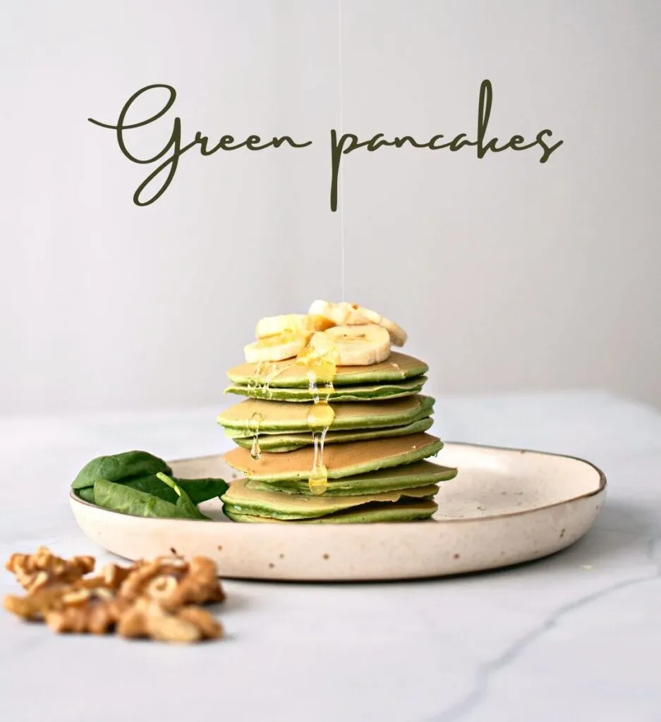 Stack of fluffy green spinach pancakes topped with sliced bananas and drizzled with honey, served on a speckled plate with fresh spinach leaves and walnuts.
